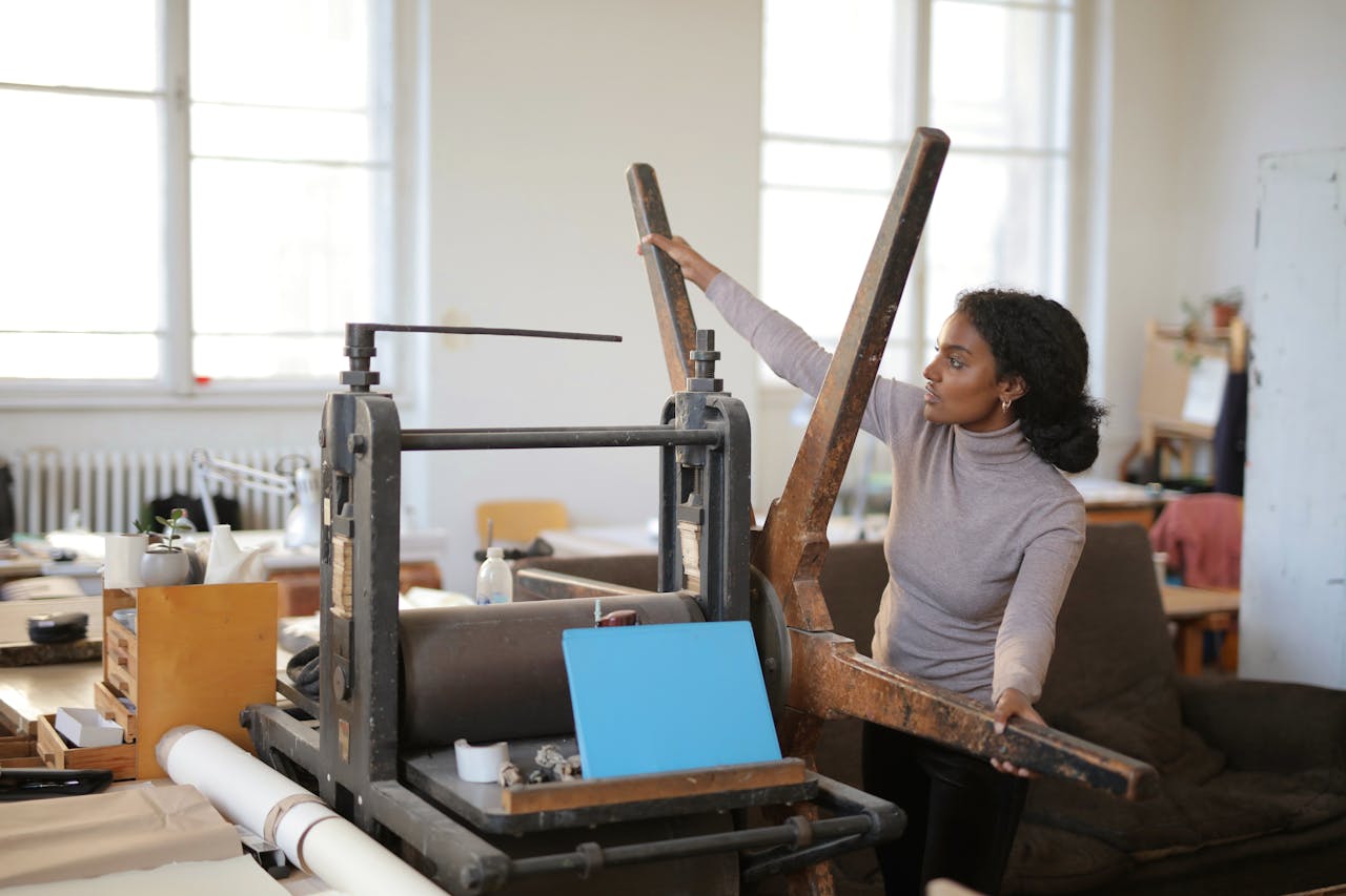 Serious African American employee taking wooden wheel of paper rolling machine while standing in workshop near paper roll in sunlight